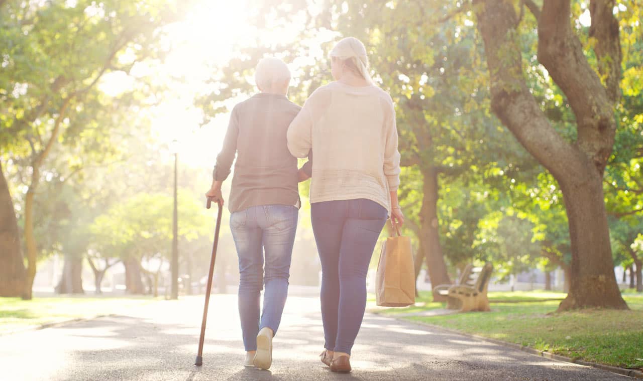 Image of daughter and elderly mother walking in sunlit park